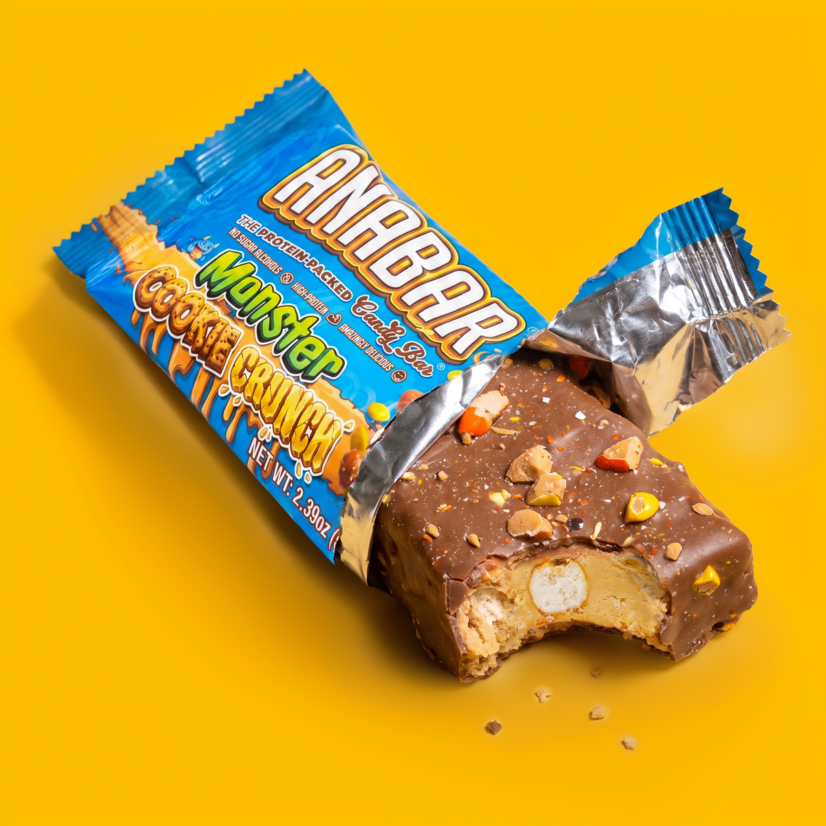 A milk chocolate-covered The Anabar Monster Cookie Crunch protein bar with a bite taken out, resting next to its partially opened blue wrapper on a yellow surface.