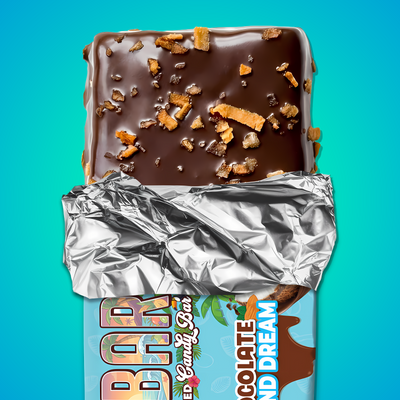 A partially unwrapped dark chocolate protein bar with caramel bits rests on shiny silver foil against a bright blue background. The wrapper reads "Coco Almond Dream" by The Anabar.