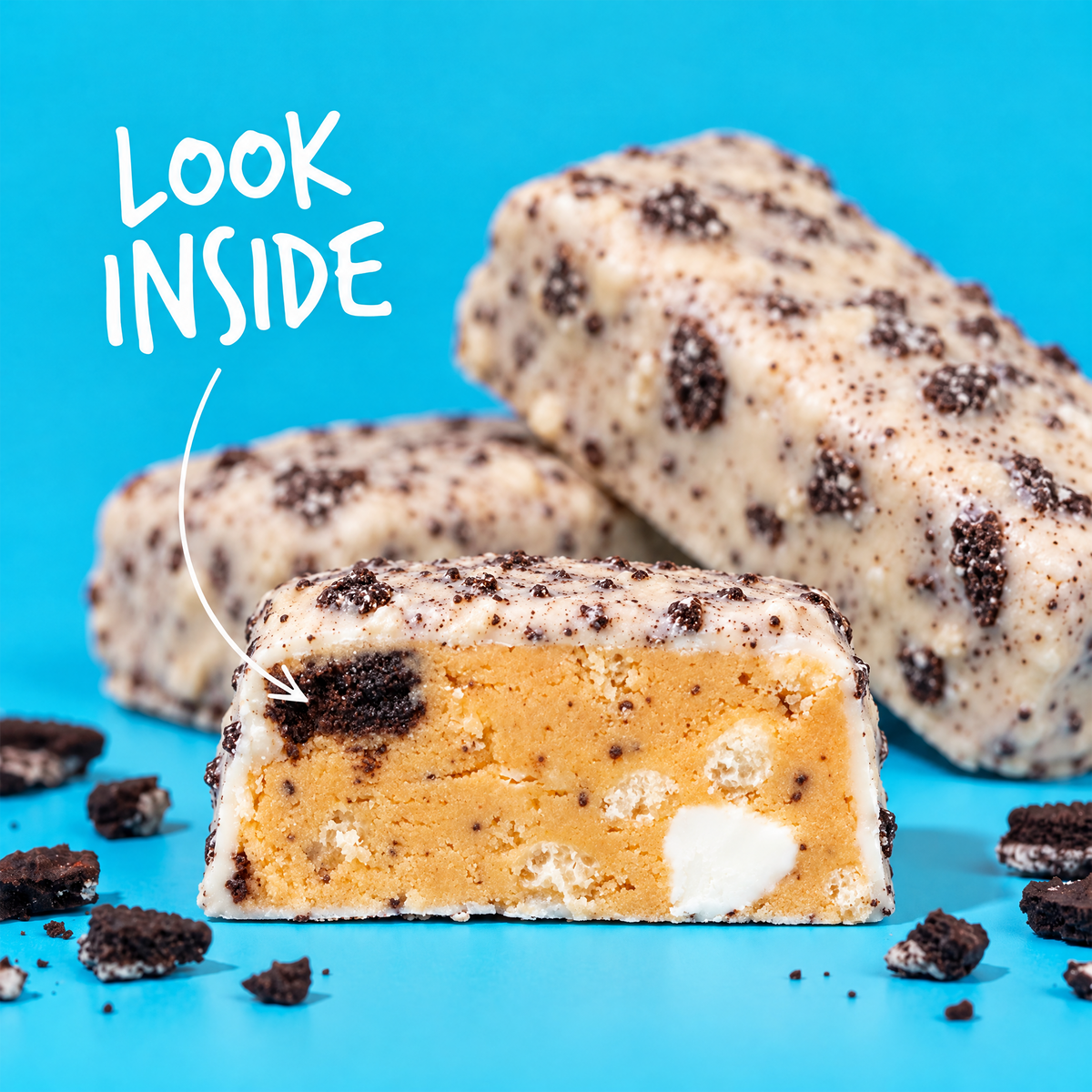 Two Cookies & Cream Anabar cookie bars against a blue background, one cut in half to show the tan interior with white inclusions and the text “LOOK INSIDE,” making for a guilt-free snack.
