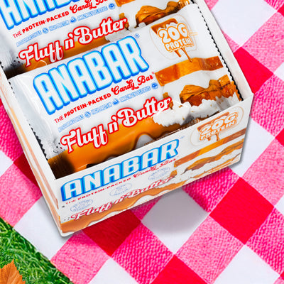 A box of The Anabar Fluff n' Butter bars rests on a red and white checkered picnic cloth, showcasing "20g protein" labels and mouthwatering images of peanut butter and marshmallow.
