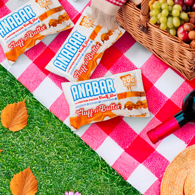 Three The Anabar Fluff n' Butter protein bars, bursting with peanut butter and marshmallow flavor, sit on a red and white picnic blanket next to grapes, wine, and autumn leaves on the green grass.