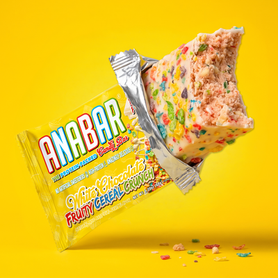 A partially unwrapped The Anabar Fruity Cereal Crunch protein bar with white icing and colorful bits, featuring a bite taken out to reveal a pink interior.