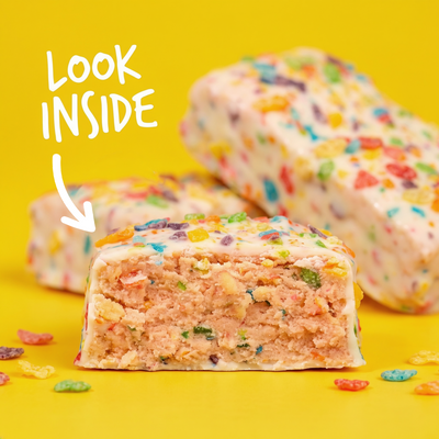 A The Anabar Fruity Cereal Crunch protein bar cut in half to show its pink interior, covered in white icing and colorful cereal pieces, with the text “LOOK INSIDE” and an arrow pointing to the cross-section.