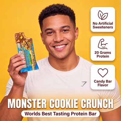 A smiling man holds a partially eaten The Anabar Monster Cookie Crunch protein bar, with icons highlighting no artificial sweeteners, 20 grams of protein, and the flavor name.