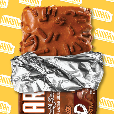 A partially unwrapped Triple Chocolate Wasted protein bar by The Anabar, featuring a milk chocolate coating, caramel layer, and chocolate sprinkles, rests on a yellow background patterned with repeating “ANABAR” logos.