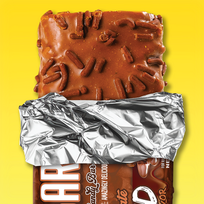 The Anabar’s Triple Chocolate Wasted protein bar, partially unwrapped from shiny silver foil, reveals its sprinkle-topped, chocolate-coated goodness against a vibrant yellow background.